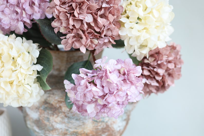 Puede incluir: Un ramo de flores de hortensia artificiales en un jarr&oacute;n r&uacute;stico. Las flores son en tonos de rosa, blanco y morado.