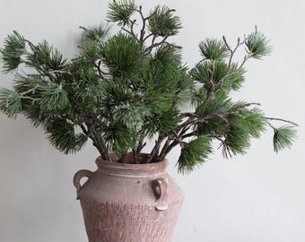May include: A rustic, terracotta-colored vase with two handles holds a collection of green pine branches. The vase has a textured surface and sits on a weathered wooden shelf. The pine branches add a natural element.