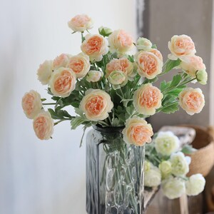 Puede incluir: Un ramo de flores artificiales de color melocot&oacute;n y blanco en un jarr&oacute;n de vidrio transparente con superficie texturizada. Las flores est&aacute;n dispuestas en un jarr&oacute;n sobre una superficie de madera.