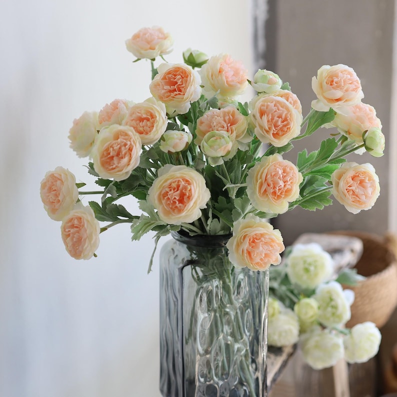 Puede incluir: Un ramo de flores artificiales de color melocot&oacute;n y blanco en un jarr&oacute;n de vidrio transparente con superficie texturizada. Las flores est&aacute;n dispuestas en un jarr&oacute;n sobre una superficie de madera.