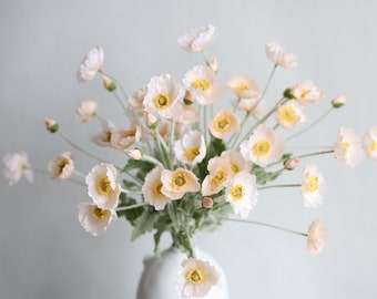 May include: A bouquet of delicate, pale pink silk poppy flowers with yellow centers arranged in a white vase.
