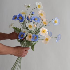 Puede incluir: Un ramo de flores de amapola de seda azul, blanco y melocot&oacute;n con tallos y hojas verdes.