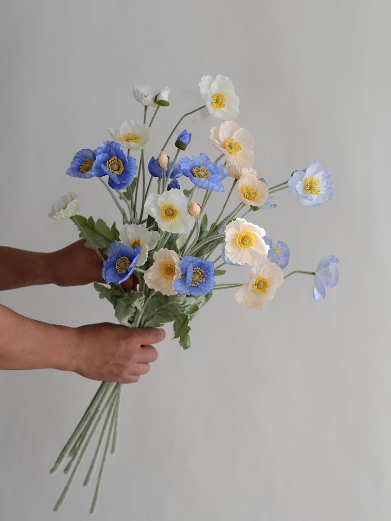 Puede incluir: Un ramo de flores de amapola de seda azul, blanco y melocot&oacute;n con tallos y hojas verdes.