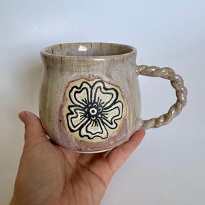 Könnte beinhalten: Eine handgefertigte Keramiktasse mit floralem Design. Die Tasse hat eine beige-violette Glasur mit einer schwarz-cremefarbenen Blume in der Mitte. Der Griff ist gedreht, und die Tasse wird in einer Hand gehalten.