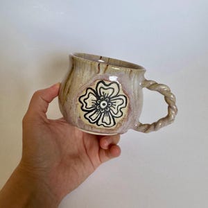 May include: A handmade ceramic mug with a floral design. The mug features a twisted handle and a beige and brown color scheme. The flower design is black and white.