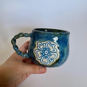 Puede incluir: Taza de cerámica hecha a mano con esmalte azul y diseño floral blanco. El asa trenzada y la altura de unos 10 cm la convierten en una pieza de cerámica única, ideal para té o café.