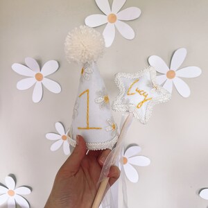 May include: White felt party hat with a white pom pom on top, decorated with yellow embroidered daisies and the number "1". A white felt star with the name "Lucy" embroidered in yellow is attached to a wooden stick with a white tulle ribbon.