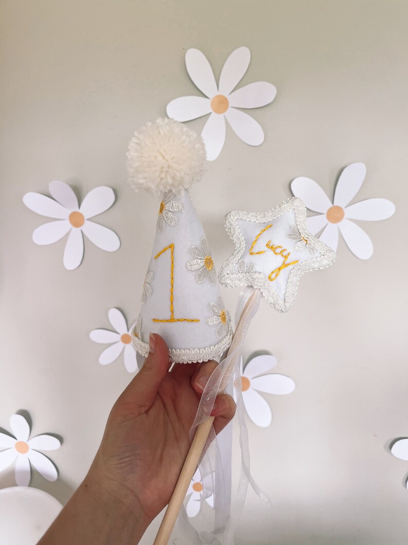May include: White felt party hat with a white pom pom on top, decorated with yellow embroidered daisies and the number "1". A white felt star with the name "Lucy" embroidered in yellow is attached to a wooden stick with a white tulle ribbon.