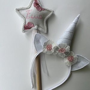 May include: A white unicorn horn headband with matching ears and a star-shaped wand. The star wand has pink embroidered butterflies and the name "Essie". The headband features white flowers with pink centers.