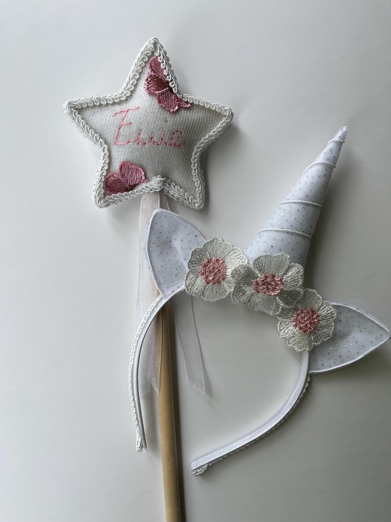 May include: A white unicorn horn headband with matching ears and a star-shaped wand. The star wand has pink embroidered butterflies and the name "Essie". The headband features white flowers with pink centers.