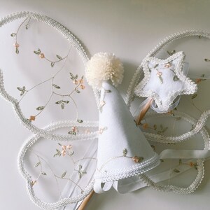 May include: A white tulle fairy costume set with a white hat, wings, and a star-shaped wand. The wings and wand are decorated with delicate floral embroidery.