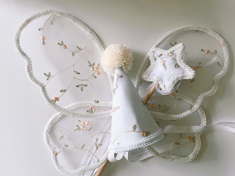 May include: A white tulle fairy costume set with a white hat, wings, and a star-shaped wand. The wings and wand are decorated with delicate floral embroidery.