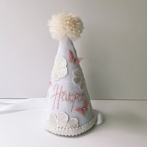 May include: White party hat with a white pom pom on top. The hat is decorated with pink butterflies and white flowers. The word "Harper" is embroidered on the front of the hat.