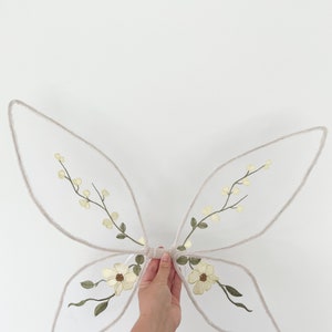 Puede incluir: Alas de hada blancas con flores amarillas y hojas verdes bordadas.