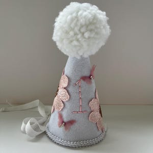 May include: A gray felt party hat with a white pom-pom on top. The hat is decorated with pink flowers, butterflies, and the number "1" embroidered in white thread.