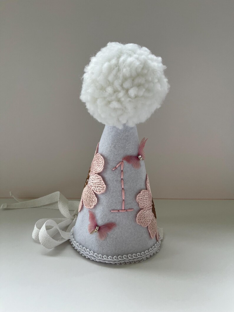 May include: A gray felt party hat with a white pom-pom on top. The hat is decorated with pink flowers, butterflies, and the number "1" embroidered in white thread.
