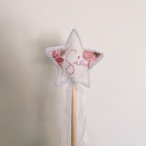 May include: A star-shaped wand with a white fabric base, embroidered with pink flowers and the name "Gia". The wand has a light blue trim, a wooden handle, and a sheer white ribbon. A whimsical accessory.