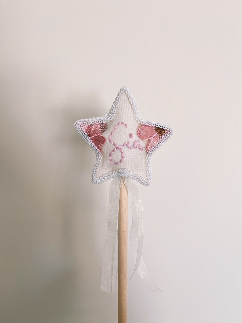 May include: A star-shaped wand with a white fabric base, embroidered with pink flowers and the name "Gia". The wand has a light blue trim, a wooden handle, and a sheer white ribbon. A whimsical accessory.