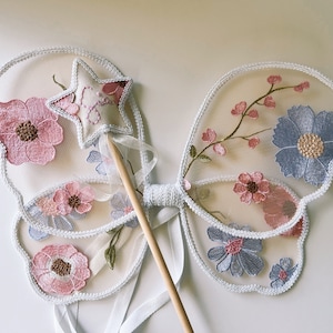 May include: Delicate fairy wings with a matching wand. The wings are made of sheer fabric, embroidered with pink and blue flowers. A star-shaped wand with a pink embroidered design and a wooden handle completes the set.