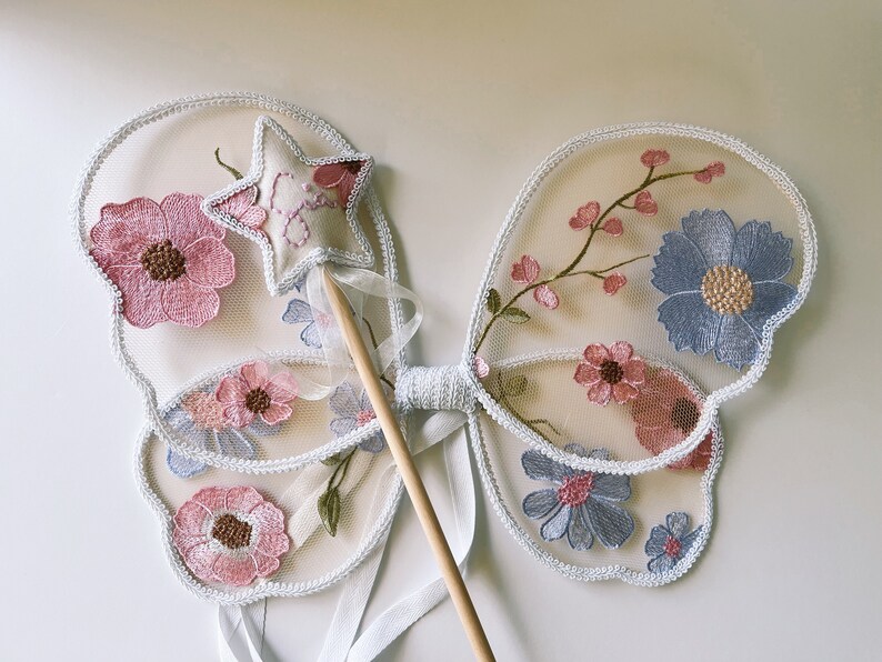 May include: Delicate fairy wings with a matching wand. The wings are made of sheer fabric, embroidered with pink and blue flowers. A star-shaped wand with a pink embroidered design and a wooden handle completes the set.