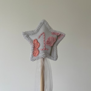 May include: A white star-shaped wand with a pink floral design and the number "1" embroidered on it. The wand has a white ribbon tied to the bottom.