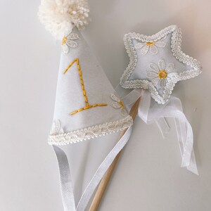 May include: White felt party hat with a white pom pom on top and a white lace trim. The hat has a yellow embroidered number "1" and white embroidered daisies. A white felt star with white lace trim and yellow embroidered daisies is attached to a wooden stick with a white ribbon.