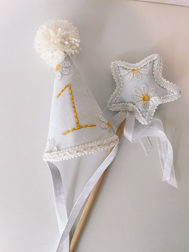 May include: White felt party hat with a white pom pom on top and a white lace trim. The hat has a yellow embroidered number "1" and white embroidered daisies. A white felt star with white lace trim and yellow embroidered daisies is attached to a wooden stick with a white ribbon.