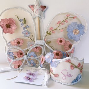 May include: A fairy costume set featuring embroidered butterfly wings, a wand, a headband, and a wrist cuff. The set includes pink and blue floral embroidery on a cream-colored mesh. A tag with the name "Samantha" is also included.
