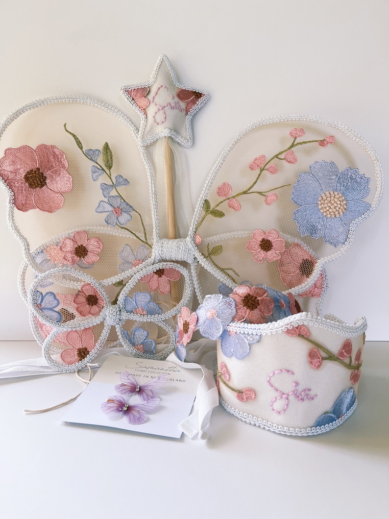 May include: A fairy costume set featuring embroidered butterfly wings, a wand, a headband, and a wrist cuff. The set includes pink and blue floral embroidery on a cream-colored mesh. A tag with the name "Samantha" is also included.