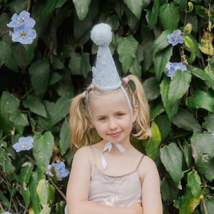 May include: A young girl wearing a white party hat with blue and silver stars and a white bow. She is wearing a light pink sleeveless dress and has her blonde hair in pigtails. She is standing in front of a green leafy background with blue flowers.