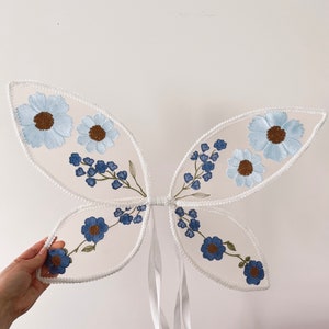 May include: White fairy wings with blue and white embroidered flowers. The wings are made of a sheer fabric and have a white ribbon tie.