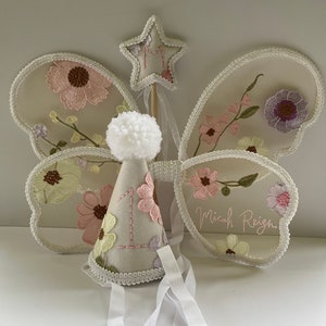 May include: A white tulle butterfly fairy wings set with floral embroidery, a white felt party hat with a number one and a white pom-pom, and a white felt star wand with the letter M embroidered on it. The wings have the name Micah Reign embroidered on them.