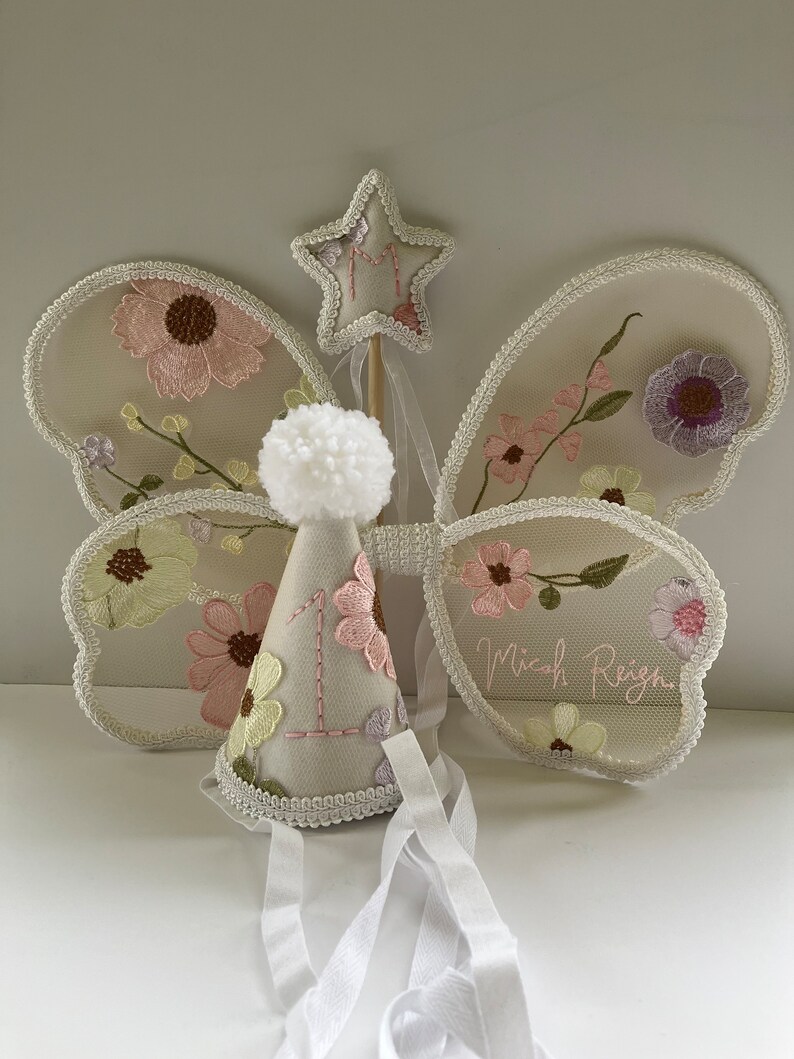 May include: A white tulle butterfly fairy wings set with floral embroidery, a white felt party hat with a number one and a white pom-pom, and a white felt star wand with the letter M embroidered on it. The wings have the name Micah Reign embroidered on them.