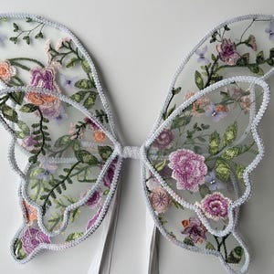Puede incluir: Un par de alas de mariposa blancas de tul con detalles florales bordados en tonos rosa, morado y verde.