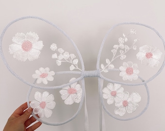 May include: White tulle fairy wings with pink flower embroidery. The wings have white satin ribbons attached.