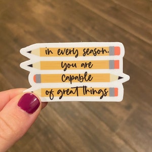 May include: A white sticker with the text "in every season. You are capable of great things" over four yellow pencil illustrations. The sticker is held by a hand with a maroon nail polish.