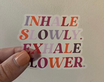 May include: A sticker with the words "INHALE SLOWLY. EXHALE LOWER." in a mix of orange, purple, and burgundy colors. The sticker is held by a hand with a light orange nail polish.