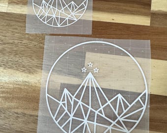 Könnte beinhalten: Zwei weiße Vinyl-Aufkleber auf transparentem Transferpapier. Beide Aufkleber zeigen eine stilisierte Bergkette mit fünf Sternen über den Gipfeln, alles in einem Kreis eingeschlossen.