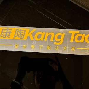 Op de afbeelding: Een rechthoekige sticker met de gele tekst "Kang Tao" en Chinese karakters. De sticker, met een transparante achtergrond, bevat ook Engelse tekst en ligt op een donkere ondergrond.