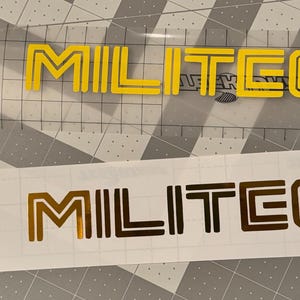May include: Two MILITECH logo decals. One is yellow on a clear background, the other is gold on a white background. Both decals feature the word "MILITECH" in a bold, sans-serif font.
