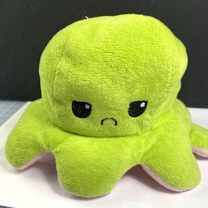 May include: A reversible plush octopus toy in shades of green and pink. One side features a grumpy face with black eyes and a downturned mouth. The other side is a happy face. The toy is made of soft, plush material.