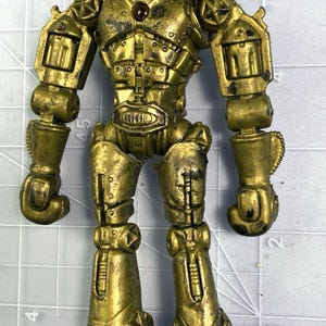 May include: A gold-coloured robot toy wearing a cowboy hat. The toy has a metallic finish with signs of wear. The robot has revolver-shaped arms and a chest with a small, red gem. The toy is approximately 15 cm tall.
