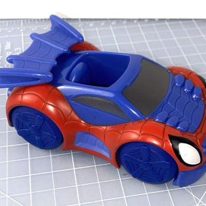 May include: A red and blue toy car with a Spider-Man theme. The car features a blue spoiler, blue wheels, and a blue cockpit. The front of the car has white eye details. The car is approximately 6 inches long.