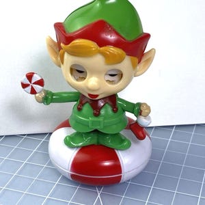 May include: A festive Christmas elf figurine. It features a green hat and outfit with red trim, and stands on a red and white striped base. The elf holds a candy cane and a peppermint candy. The figurine has a smiling face.