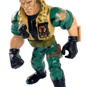 May include: A vintage action figure of a muscular man in a military-style outfit. The figure wears a camouflage jacket, green pants, and black gloves and boots. The figure has gray hair and a stern expression.