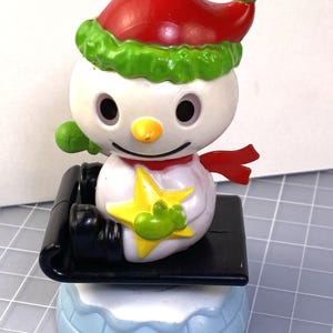 May include: A snowman figure wearing a red hat with green trim and a red scarf. The snowman has a yellow nose, black eyes, and a smiling mouth. It sits on a black sled, on a light blue base with white trim.