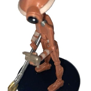May include: A brown and tan figurine of a robot-like character with a large head and a tool. The figure is standing on a black circular base. The tool has a silver shaft and a gold-colored end.