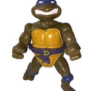 May include: A vintage action figure of Leonardo from Teenage Mutant Ninja Turtles. The figure is brown with a yellow plastron, purple belt, and blue accents. It has a wide, toothy grin and is posed in a dynamic stance.