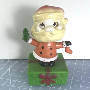 May include: A vintage Santa Claus figurine with a cream-colored head and a red and orange coat. The figurine is holding a green Christmas tree and is standing on a green gift box with a red bow. The figurine has black boots and gloves.
