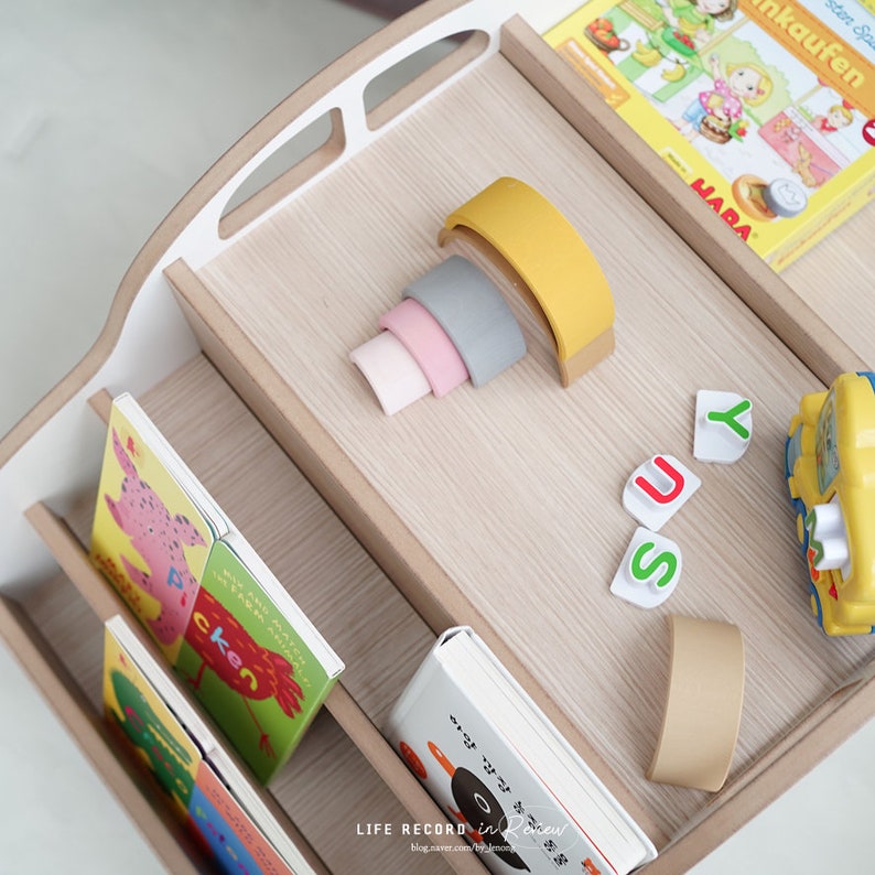 May include: A wooden bookshelf with three shelves, filled with colorful children's books. There are also a few colorful stacking rings, a yellow toy bus, and letter blocks that spell "JUSY" on the top shelf.